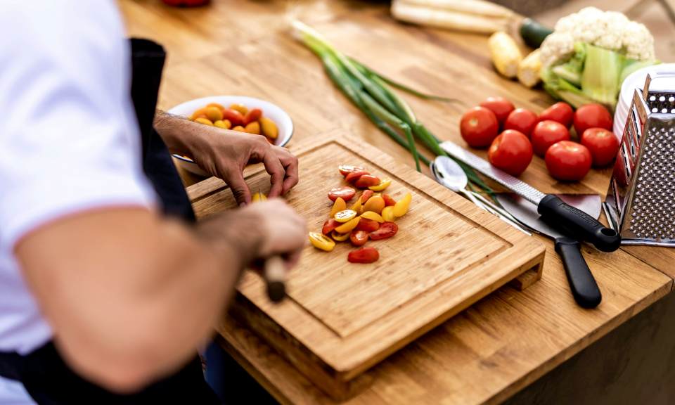 Alta cocina vegetal – Mindful cooking en la Hospedería de la Corte Manos de un chef cortando tomates y verduras frescas sobre una tabla de madera, representando la alta cocina vegetal y el mindful cooking del Retiro de Perfumería Natural y Mindful Eating en la Hospedería de la Corte, Sierra de Aracena.