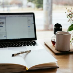 Mesa de trabajo con cuaderno, bolígrafo, taza de café y portátil, símbolo del proceso creativo de escritura en la Hospedería de la Corte.